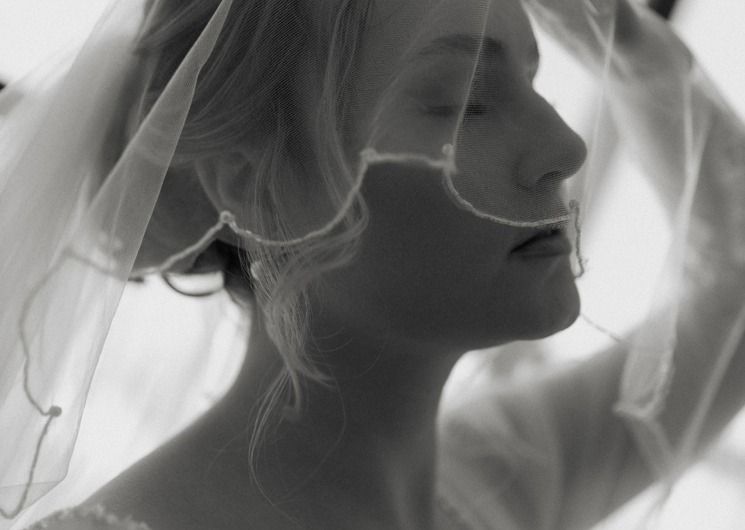 Black and white wedding portrait of a bride lifting her veil in soft window light