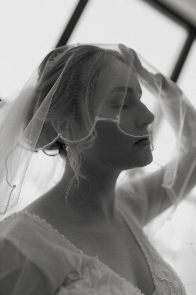 Black and white wedding portrait of a bride lifting her veil in soft window light
