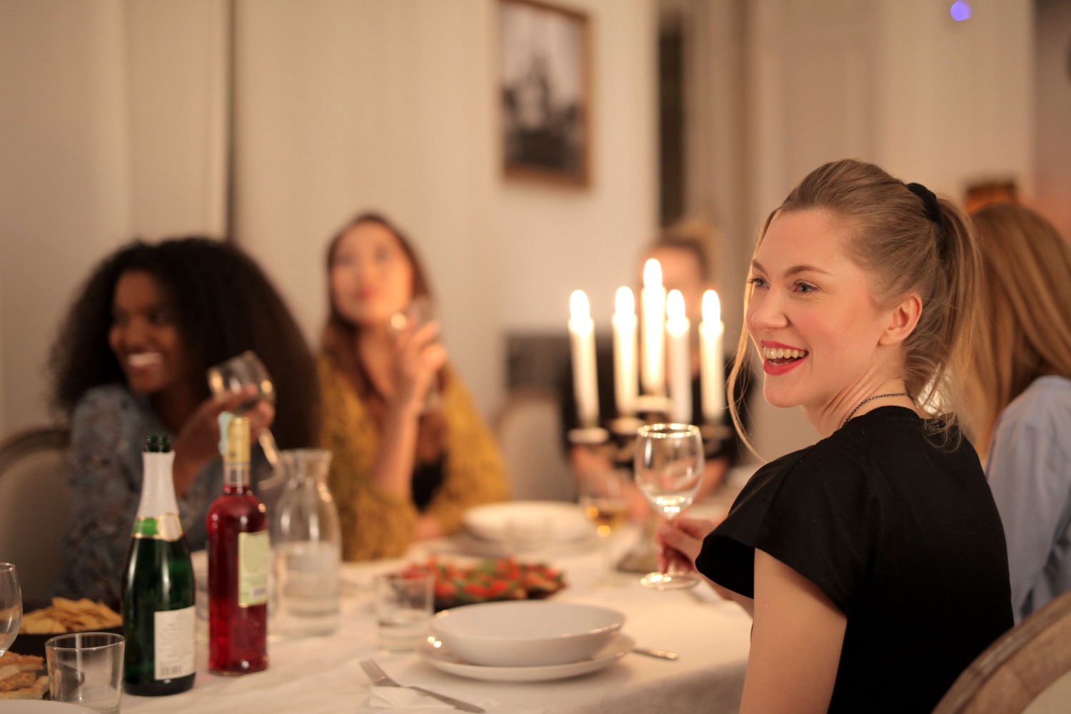 Group of friends laughing around a candlelit dinner table with wine glasses and sharing plates