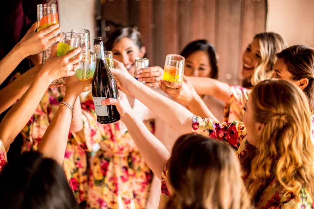 Group of women in floral robes raising colourful glasses and a bottle of prosecco together in a toast